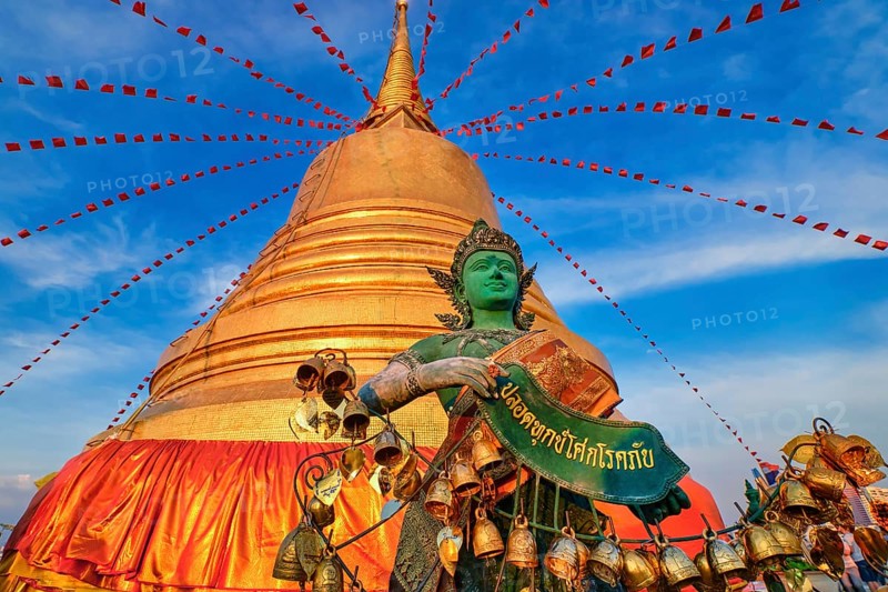 Statue of green-skinned Thai angel holding roll with message in Thai stand in front of big golden stupa in sunlight and at blue sky with clouds in Buddhist temple. Decorations with bells