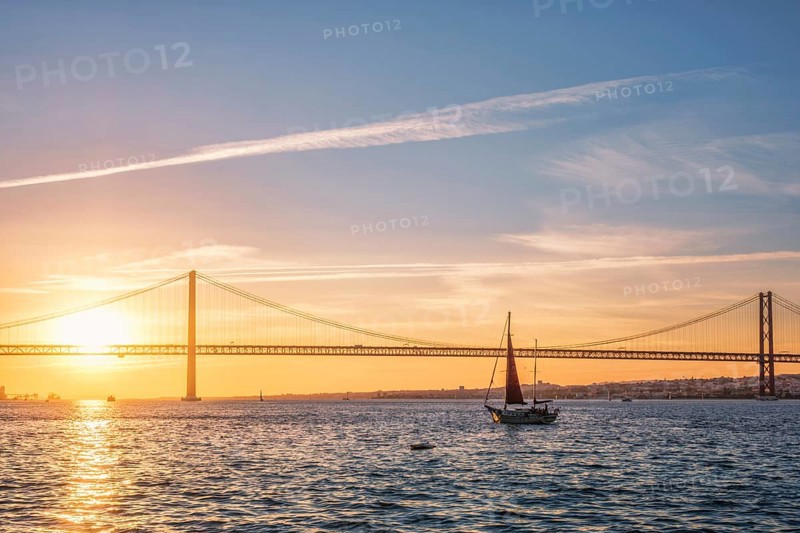 View of 25 de Abril Bridge famous tourist landmark of Lisbon connecting Lisboa and Almada over Tagus river with tourist yacht silhouette at sunset. Lisbon