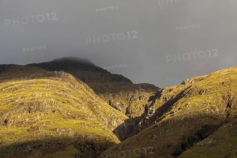 Rain cloud forming thick fog descending from steep mountainside bading in evening light at sunset in Glen Coe