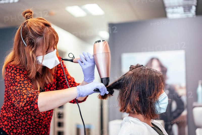 Blonde hairdresser with mask and gloves straightening client's brown hair with blow dryer. Reopening with security measures for hairdressers in the Covid-19 pandemic