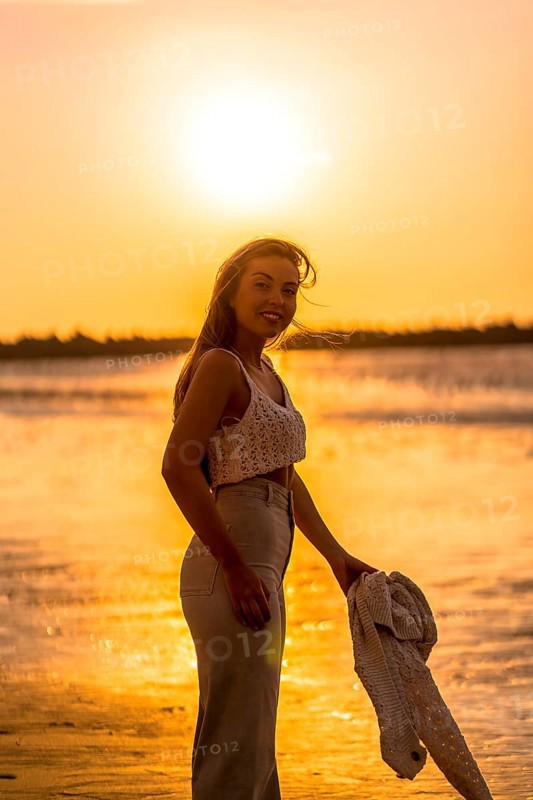 Summer lifestyle. A young blonde Caucasian woman in a white short wool sweater on a beach sunset. With eyes closed and enjoying the sea wind