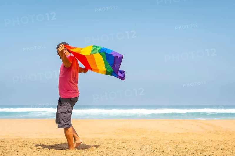 A gay person with a pink t-shirt and black cap with the LGBT flag on a beach