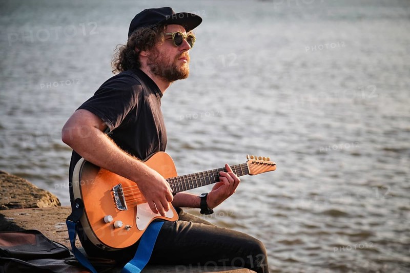 Hipster street musician in black playing electric guitar in the street sitting on pier embankment on sunset