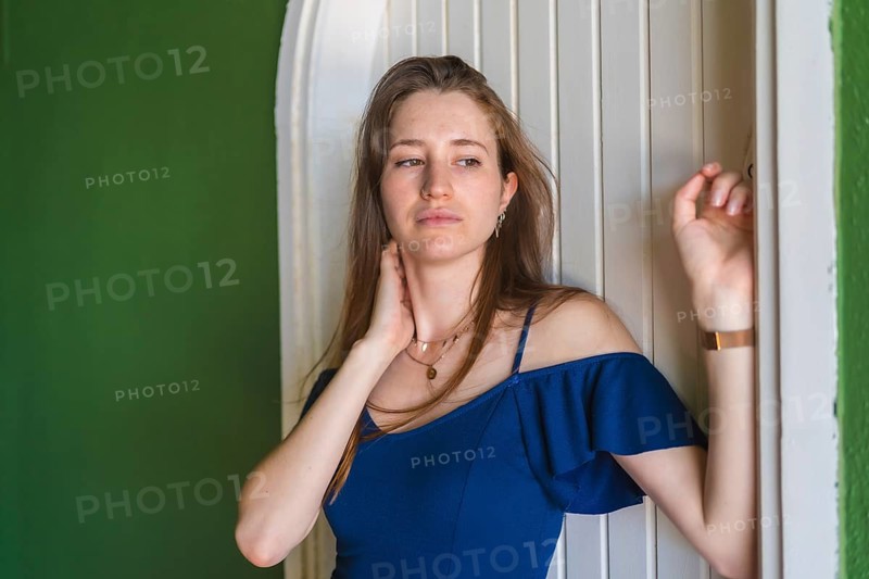 A young redhead Caucasian girl smiling in a blue dress next to a white door of a green house