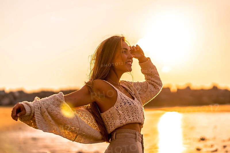 Summer lifestyle. A young blonde Caucasian woman in a white short wool sweater on a beach sunset. With open arms by the sea