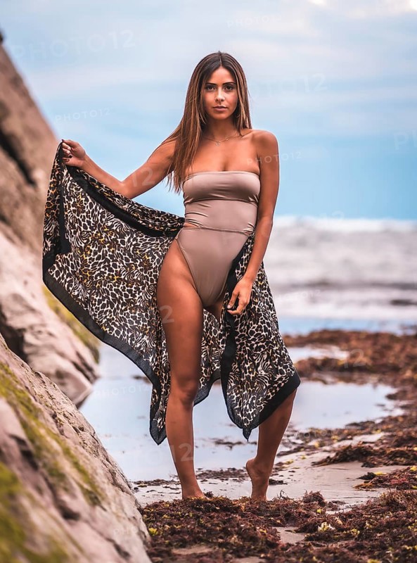 A dark-haired caucasian woman in a brown swimsuit and sarong on a natural background next to rocks and sea. In the summer sunset
