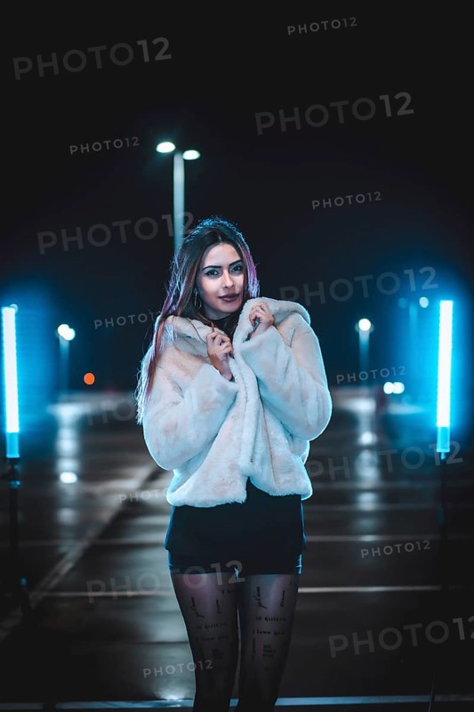 Young Caucasian brunette with pink wool jacket in an empty car park illuminated with blue light. Night urban winter session in the city