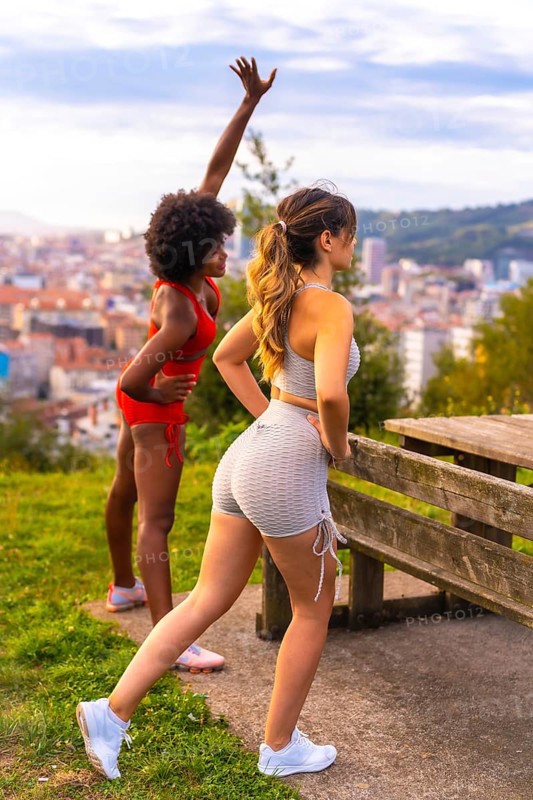 Caucasian blonde girl and dark-skinned girl with afro hair doing stretching before starting sports in the park. Healthy life