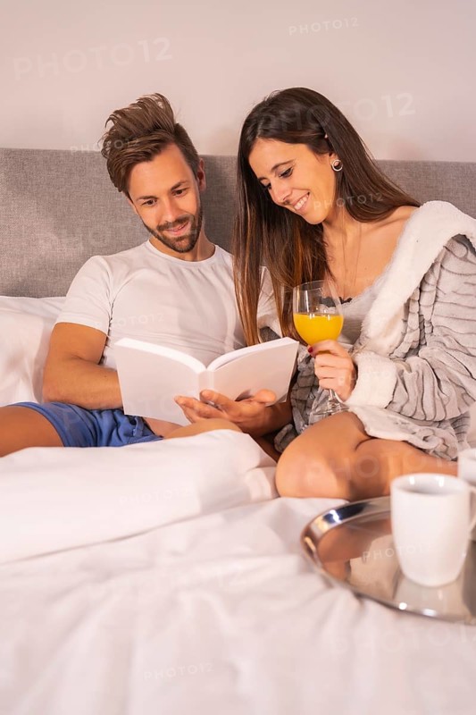 A couple in pajamas reading a book in the breakfast of coffee and orange juice in the hotel bed