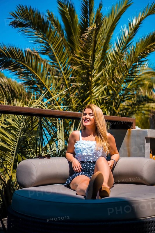 Summer lifestyle of a young blonde Caucasian woman in a white flowery top enjoying the summer on a terrace with palm trees. Sitting on a sofa on a terrace sunbathing and smiling