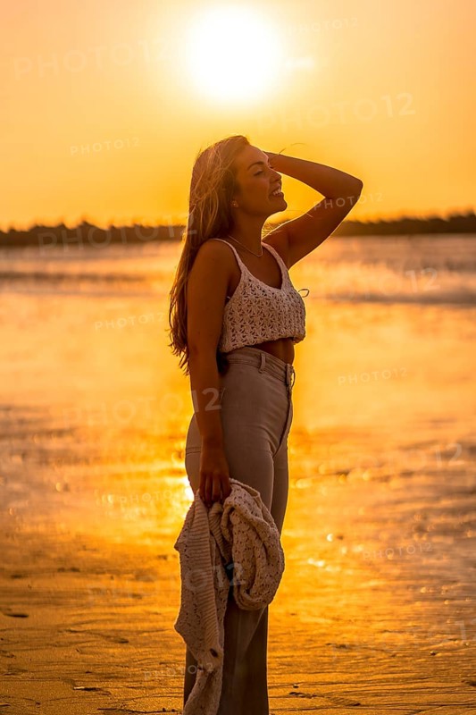 Summer lifestyle. A young blonde Caucasian woman in a white short wool sweater on a beach sunset. With eyes closed and enjoying the sea wind