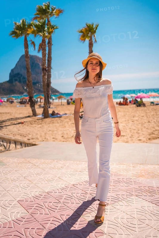 A Caucasian redhead dressed in white and with a straw hat walking along the beach of Calpe in summer