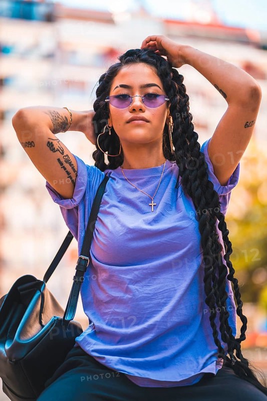 Portrait of young dark-skinned woman with long braids and purple glasses sitting on a bench in the park