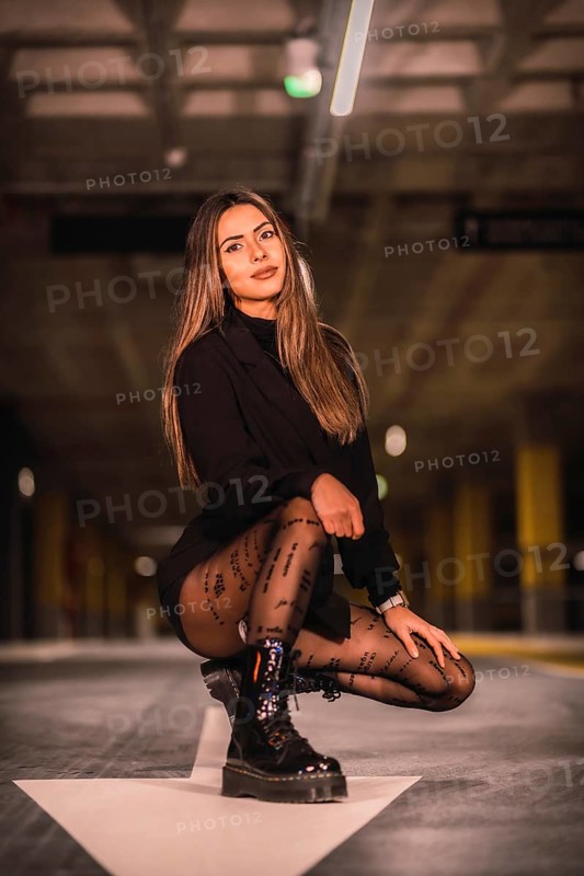 Caucasian brunette model with black jacket posing on the white arrow of the ground in an empty underground car park. Night urban session in the city
