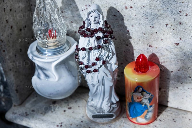 Kitschy electric battery-powered candles next to a small rosary-wrapped figure of Mary on an urn grave in a cemetery