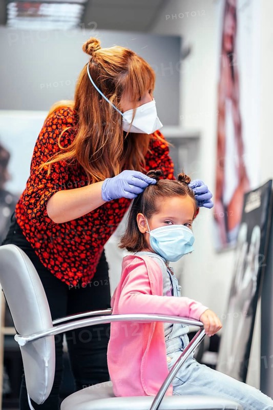 Hairdresser with mask and gloves finishing the pigtails to a girl. Reopening with security measures for hairdressers in the Covid-19 pandemic