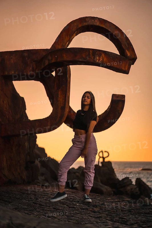 A young pretty brunette Latina with long straight hair in a short black T-shirt and pink pants. Perched next to a famous sculpture of Saint Sebatian called Peine del Viento