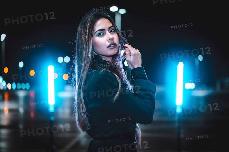 Caucasian brunette in pink wool jacket in an empty car park illuminated with blue light. Night urban session in the city