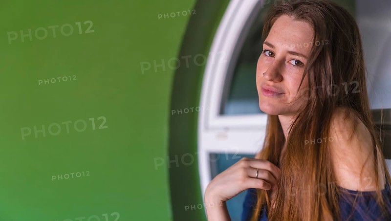 A smiling young pretty redhead Caucasian woman sitting in a blue dress next to a white sale of a green house