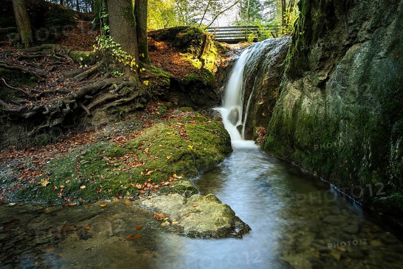 A small waterfall near the Gerats waterfall with moss-covered rocks and tree roots. The sun is shining in the background. Gerats