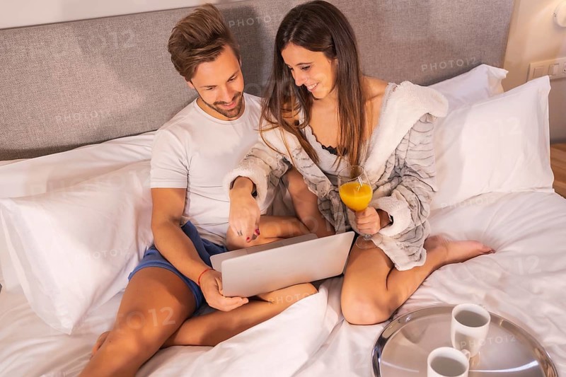 A couple in pajamas looking for hotels or trips from the computer in the breakfast in the hotel bed