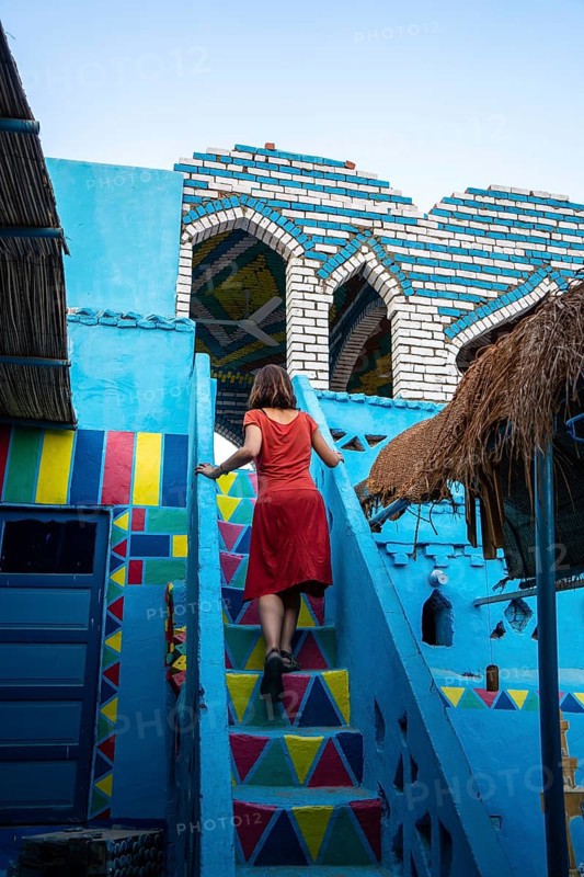 A young woman in a red dress climbing some colorful stairs that go up to a beautiful terrace of a traditional blue house in a Nubian village next to the Nile river and near the city Aswan. Egypt