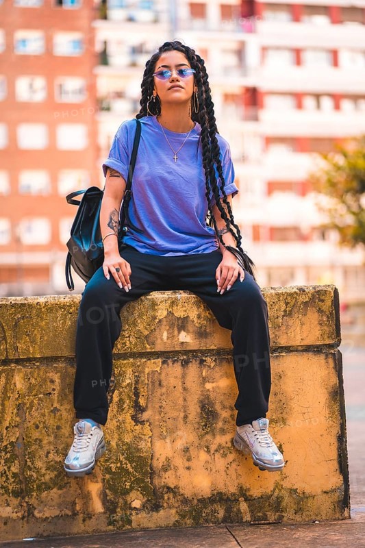 Dark-skinned young woman with long braids and purple glasses sitting on a bench in the city