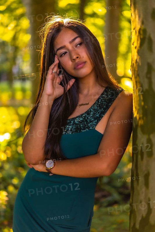 A young brunette Latina with long straight hair leaning against a tree in a green dress. Portrait with a sweet and sensual look