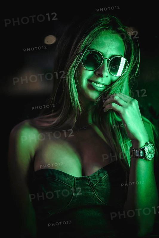 Caucasian brunette in a black dress illuminated by green led light reflected in black crystals. Urban night photography. Girl with sunglasses and saying quiet