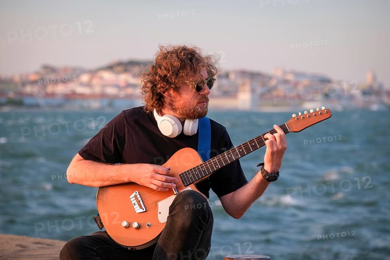 Hipster street musician in black playing electric guitar in the street on sunset on embankment with Lisbon in background. Portugal