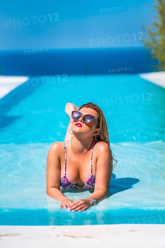 Summer lifestyle in an infinity pool with a young blonde Caucasian woman in a pink and purple bikini with sunglasses. Lying in the pool water with the ocean in the background