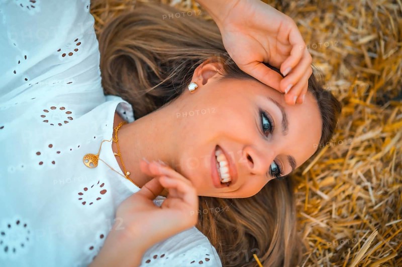 A young blonde Caucasian woman in a white dress in a field of dry straw atop a haystack