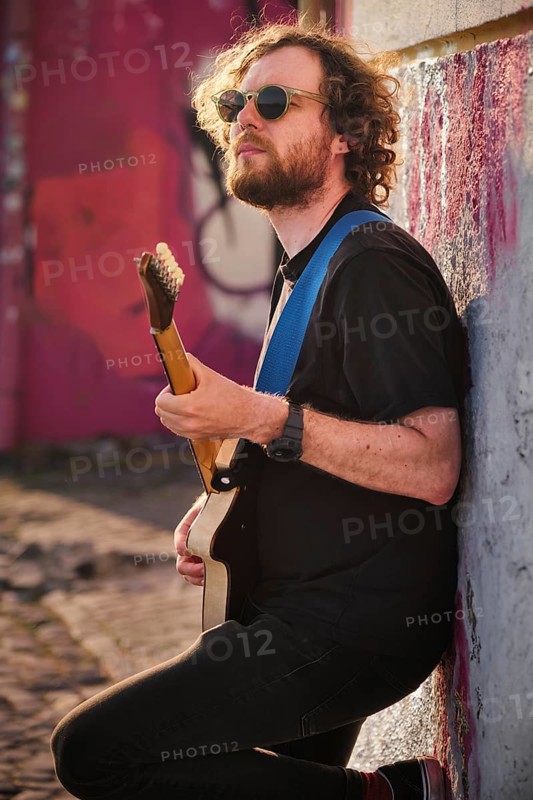 Hipster street musician in black playing electric guitar in the street on sunset leaning on a wall