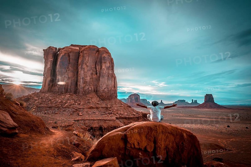 A boy in a white T-shirt sitting on a stone with his arms up in the sunset inside Monument Valley National Park. Utah