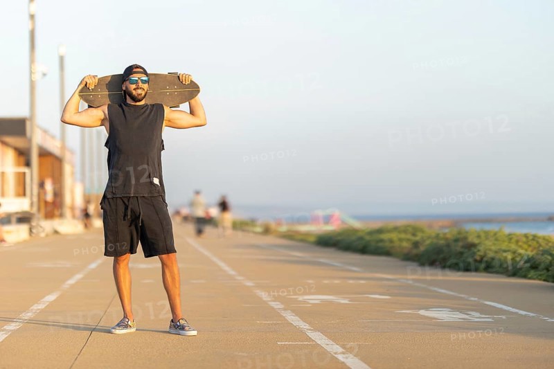 A man standing on the bike path holding a skateboard on his shoulders. Mid shot
