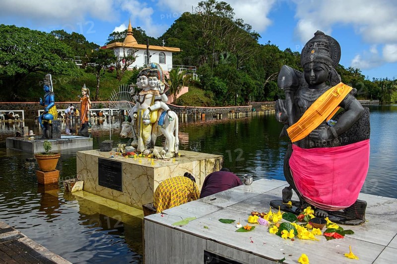 Right Hindu deity statue figure sculpture of goddess deity of Lord Muruga left beside Dattatreya triune figure Brahma Vishnu beside Lord Shiva in background woman Shakti of Shiva Hindu mother goddess mother of Ganesha in lake sacred Hindu lake Ganga Talao at religious site largest Hindu sanctuary sanctuary for religion Hinduism outside India for devout Hindu Hindus