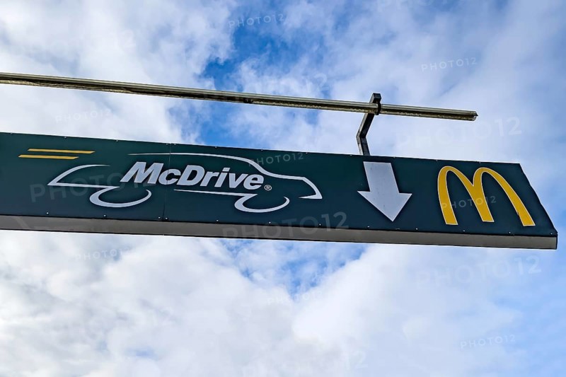 Sign mounted at height on pole with crossbar with white lettering McDrive and logo yellow M of McDonald's above entrance from drive in to fast food restaurant