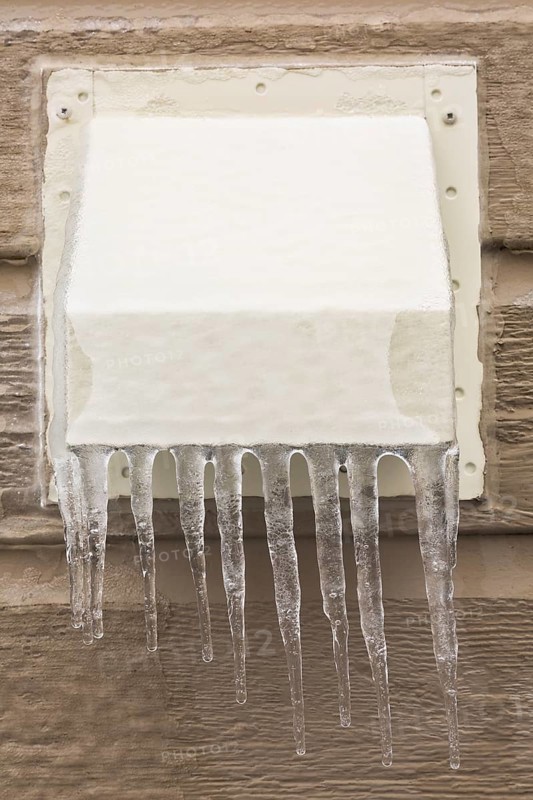 Metal exhaust vent covered with ice and icicles on tan clapboard wall of house