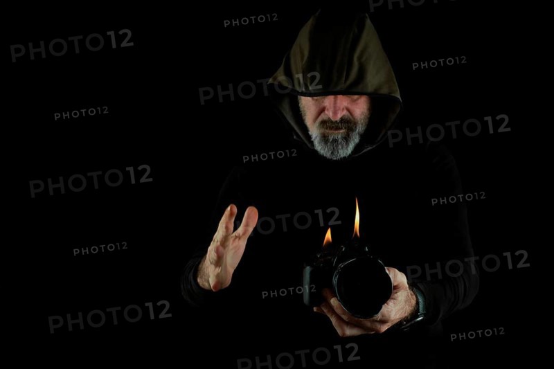 Man dressed in black with hood holding a burning reflex camera on a black background