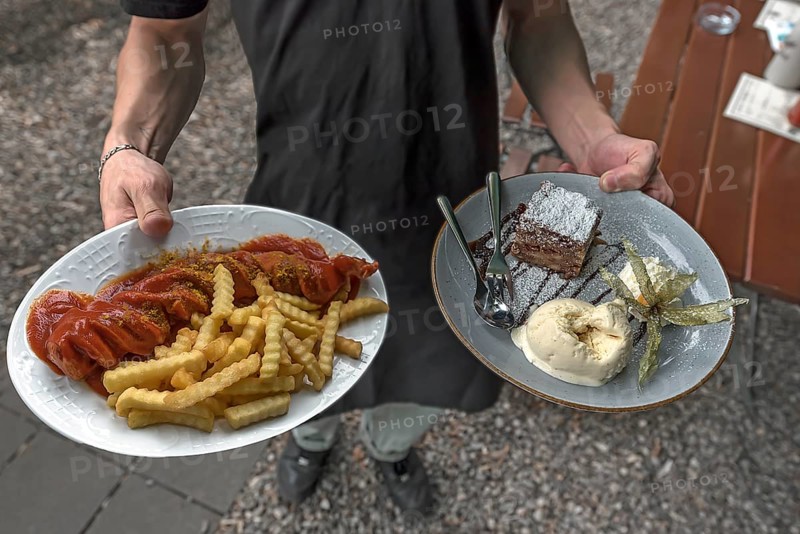 A waiter serves currywurst with fries and apple strudel with ice cream in a garden restaurant