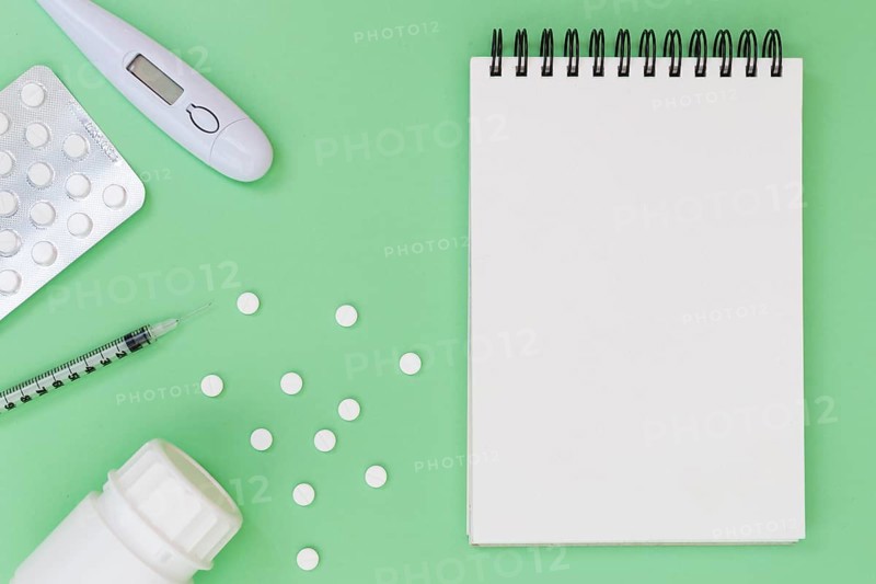 Pills thermometer syringe plastic bottle near spiral notepad green background