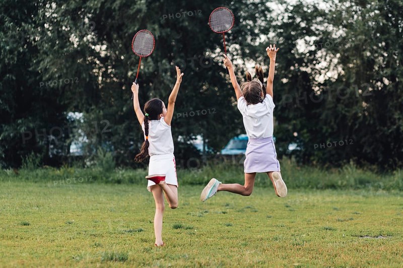 Rear view two girls jumping park holding badminton