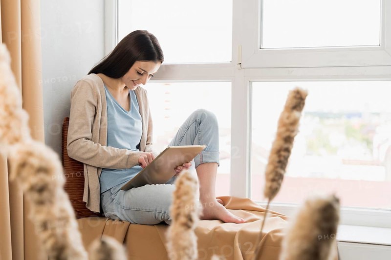 Side view woman sitting window working tablet