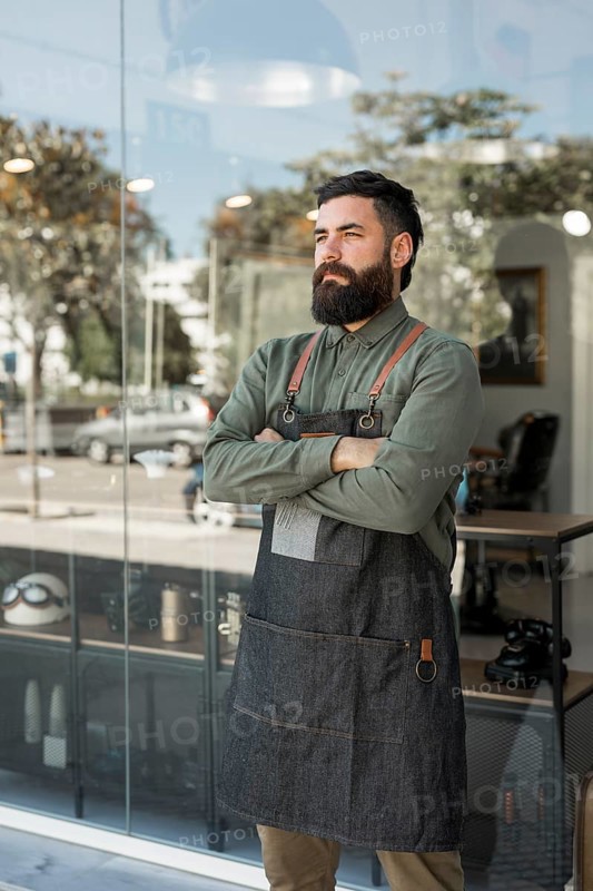 Bearded serious hairdresser standing outside near barbershop