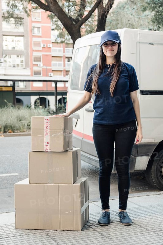 Portrait young female courier standing with stacked cardboard boxes