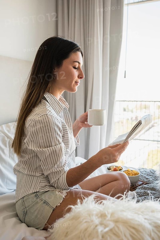 Young woman sitting bed reading newspaper while drinking coffee