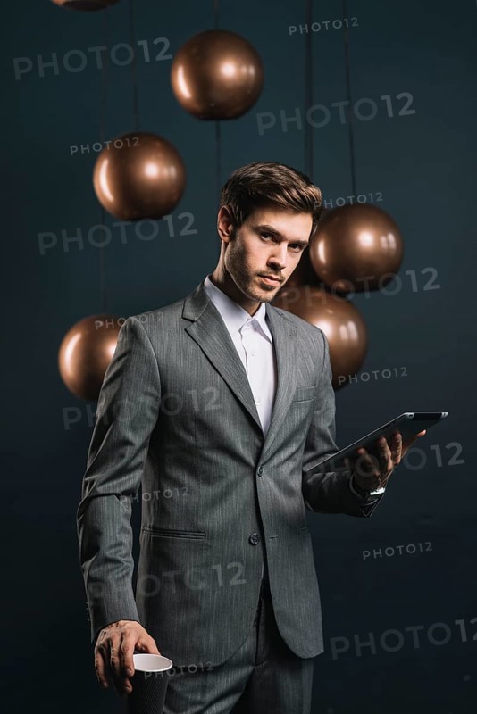 Confident young man holding disposable cup digital tablet against dark background