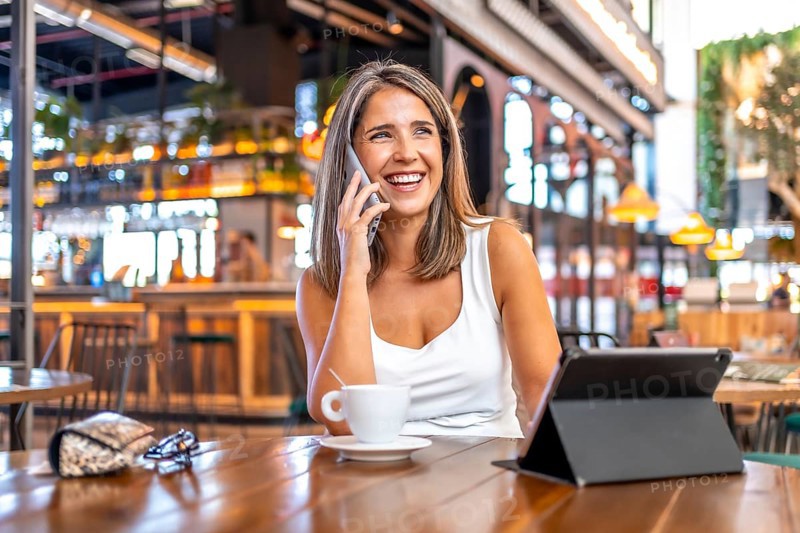 Distracted businesswoman talking to the phone in a cafeteria sitting with a digital tablet and a coffee cup
