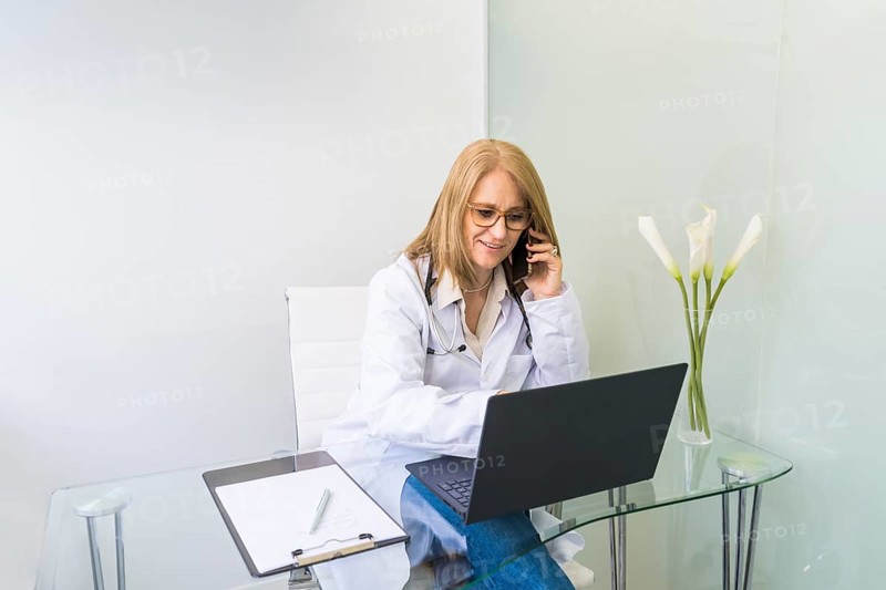 Smiling female doctor talking on the phone to a patient while looking at her medical records on laptop in medical office