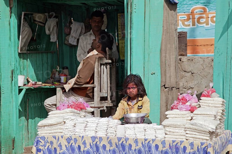 Hairdresser cutting hair in barbershop and child behind display selling goods in Govardhan